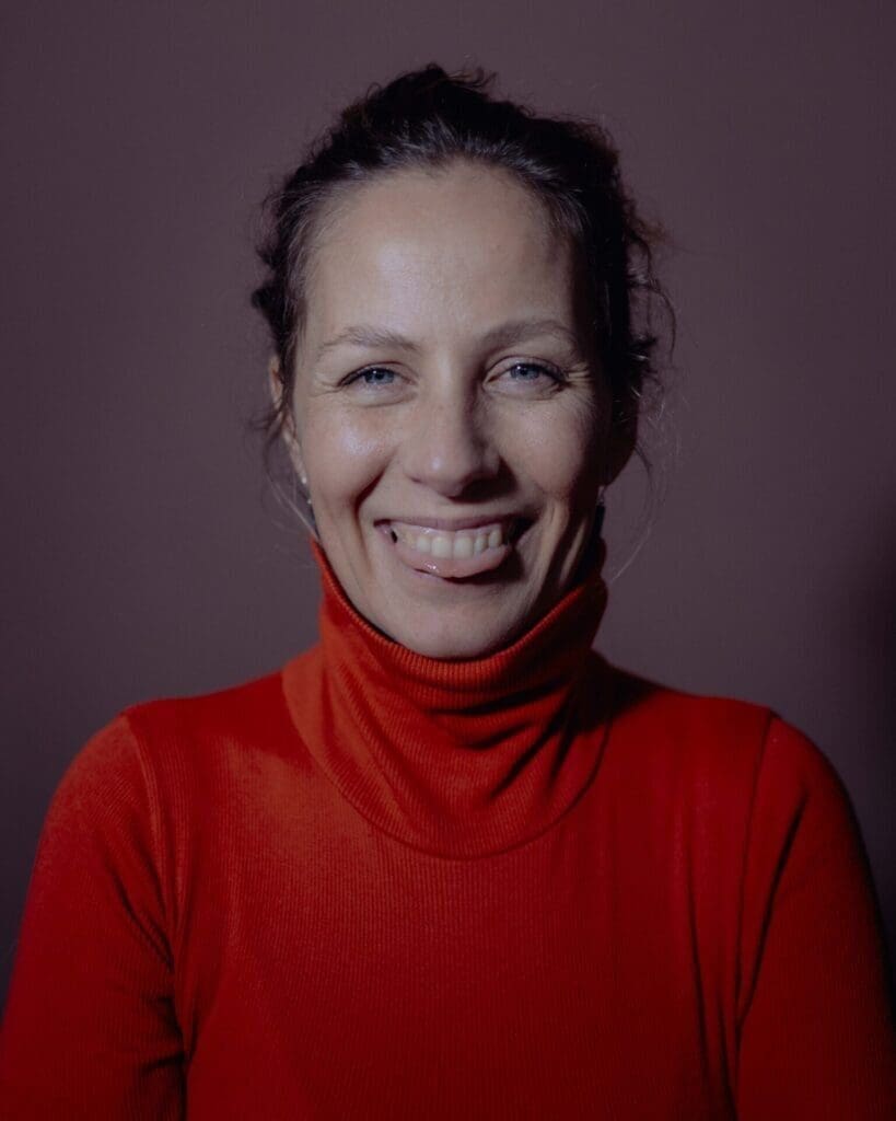 khandiz Khandiz, a white woman with her hair loosly pulled back. She's wearing a red polo neck and standing against a dark pink background. She's cheekly pulling a tongue at the camera